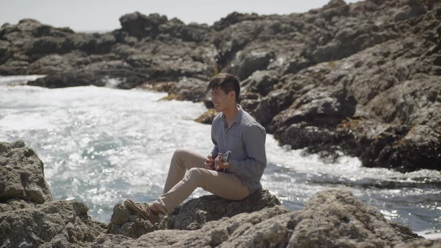 A man sitting on a rocky shore playing the ukulele