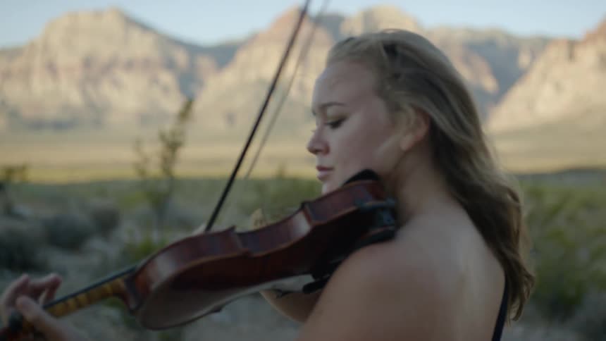 A woman with blonde hair stands outdoors before a mountain range while playing the violin