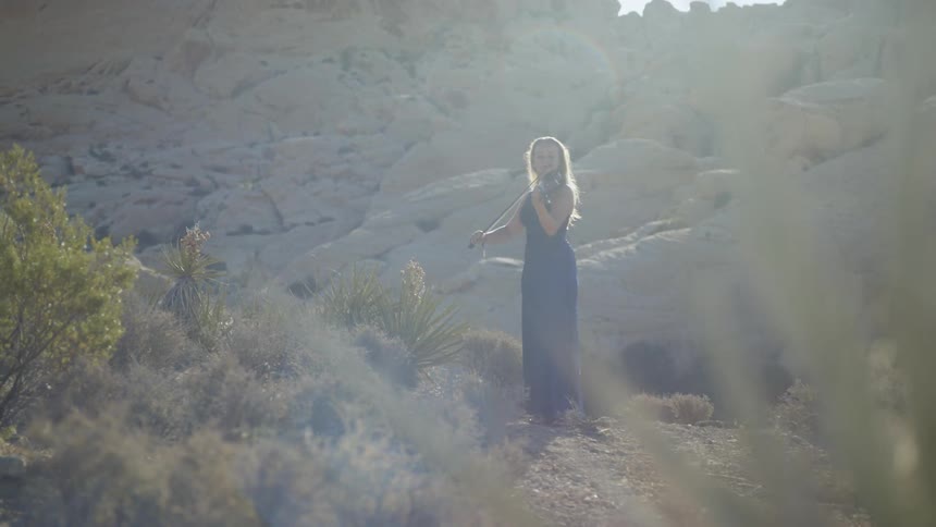 Blonde woman wearing a blue dress plays on the violin in the canyon on a sunny day