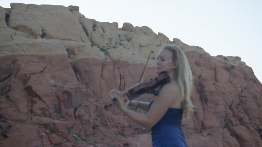A woman in a blue dress plays her violin near a rock formation