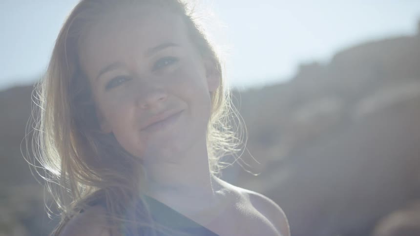 A blonde haired woman standing outdoors in the direct sunlight smiles warmly