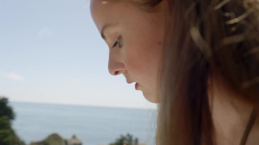 A woman stands above a mountain side coast line with a pencil and notepad