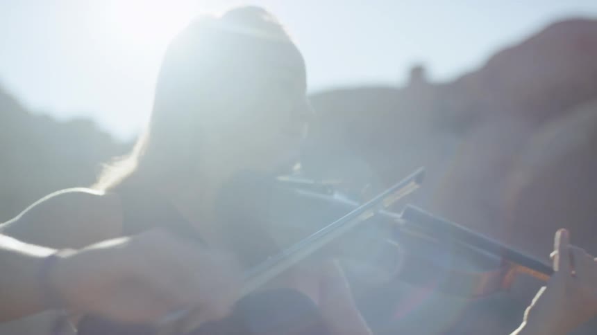 A blonde haired woman standing outside in the sunlight plays a violin