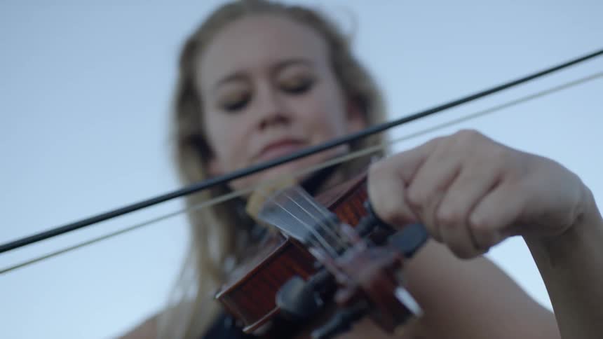 Female rests a violin under her chin and glides the bow across the strings as she turns a peg on the neck