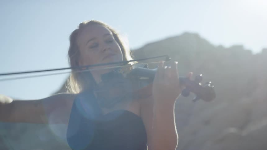 A woman is playing the violin outdoors surrounded by rocky terrain