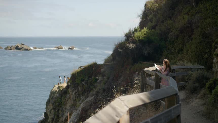 A woman is holding a notebook as she stands against a railing near the ocean
