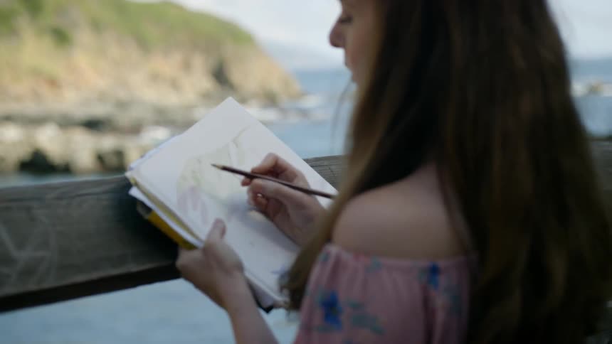 A woman is standing near the coast drawing in a notebook
