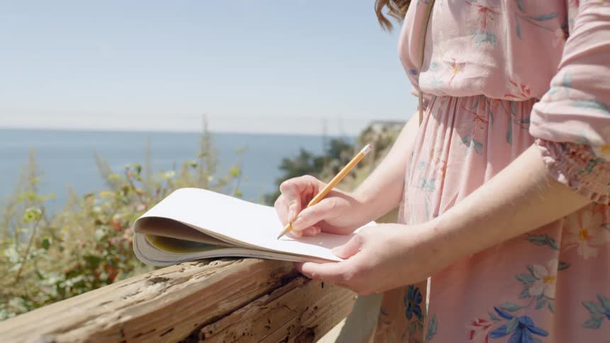 A woman stands on a deck writing on paper near the ocean