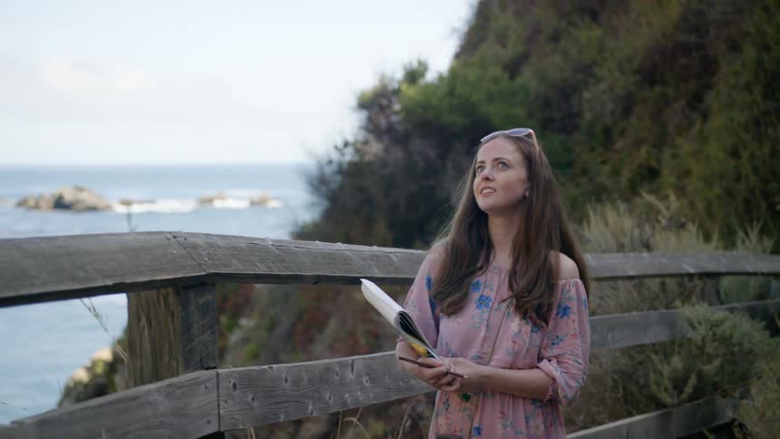 A woman walks across a small bridge by the water with a notepad in her hands