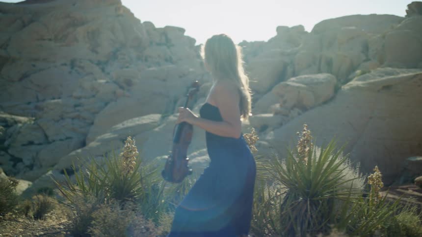 A woman holding a violin is walking past hills in the desert