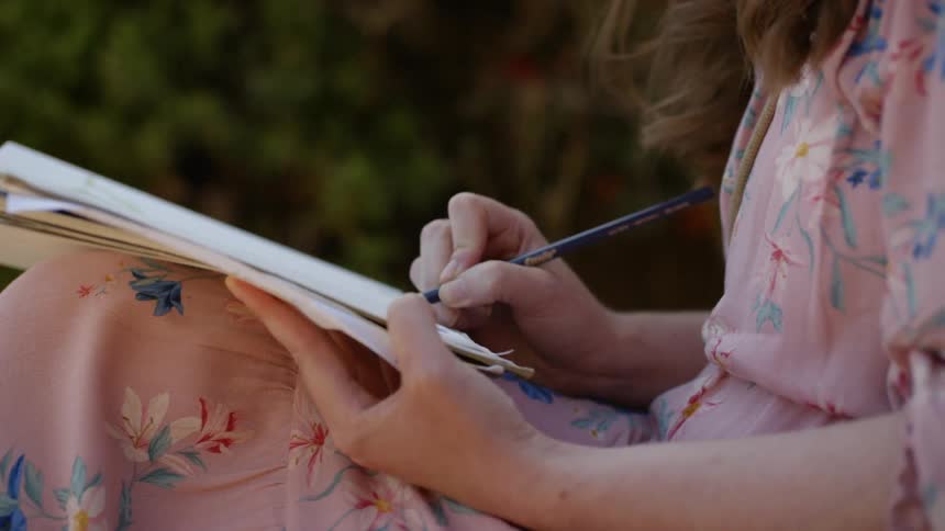 A woman wearing a floral print dress sits outside and sketches with a pencil
