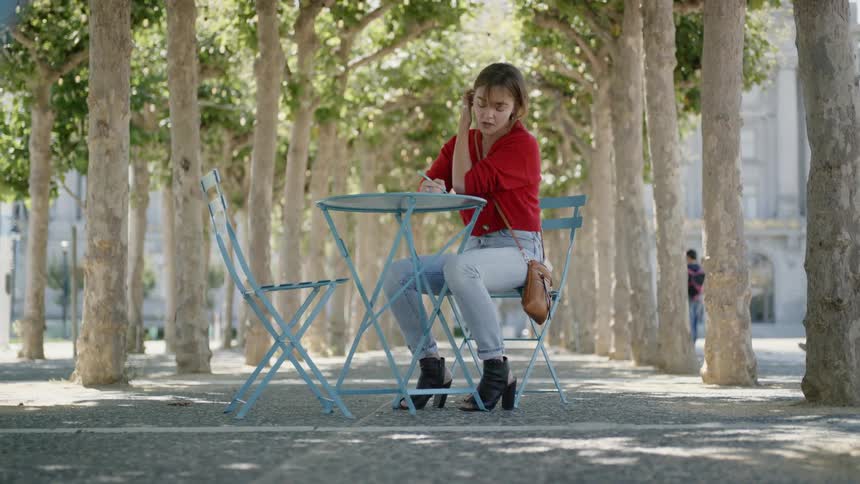 Woman sitting at an outdoor table in between a path of trees writing in book