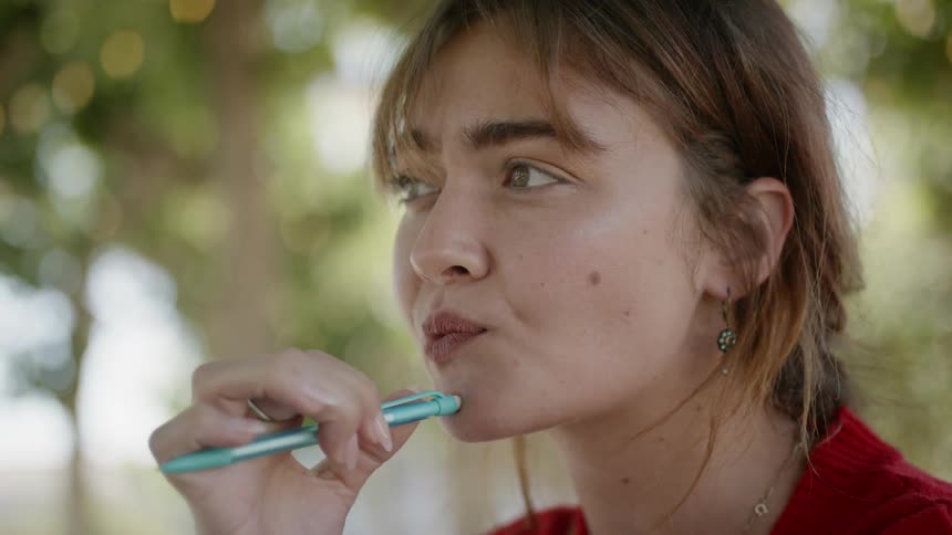 A young woman pushes a pen against her chin and thinks and then writes in a book