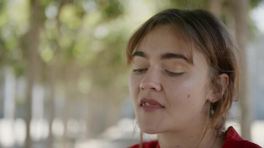 A woman sitting under a row of trees in a park removes her glasses while thinking