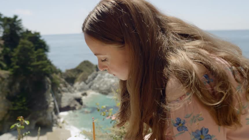 A woman writes in a notebook outside near a beach