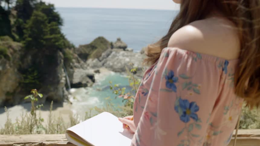 A woman stands on a pathway looking to the ocean water with a sketchpad and pencil