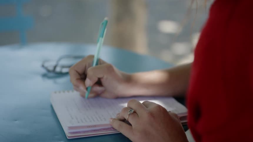 A woman sits at an outdoor table writing in a notebook