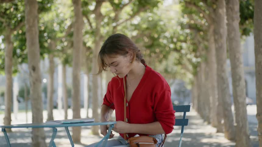 A young woman sits at a table outdoorswhile cleaning her glasses and putting them back on