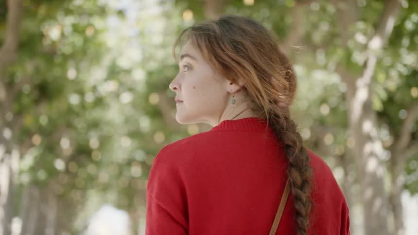 A woman with a red sweater walking under trees with sunlight shining on her