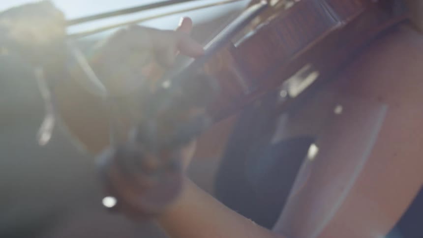 A white female with long blonde hair is playing a violin outside with the sun in the sky behind her