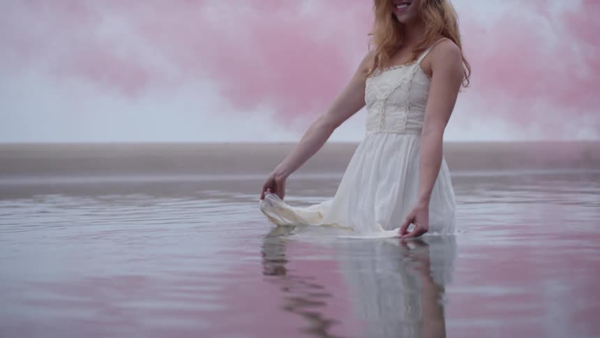 The woman in the white dress standing in water and smiling with pink smoke and the sky in the background