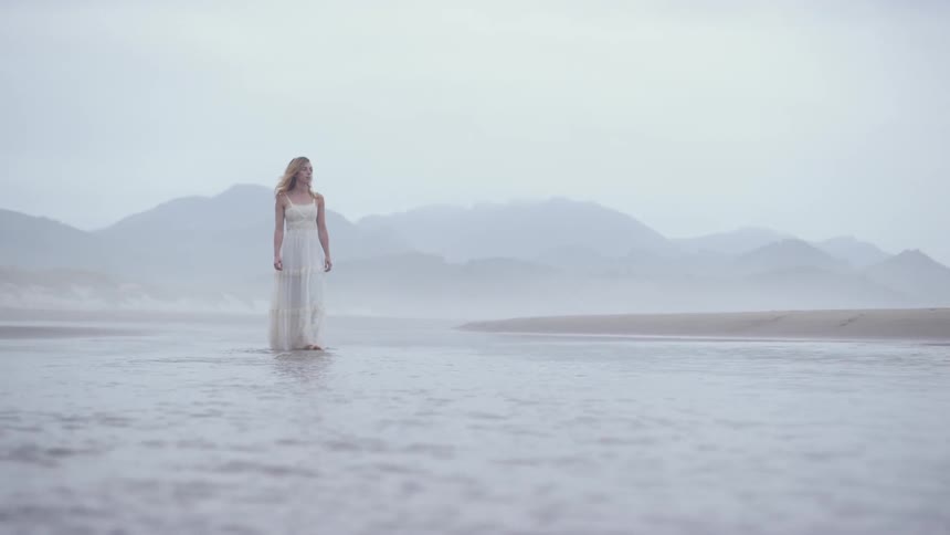 Blonde woman wearing a white dress walks on the beach on a cloudy day
