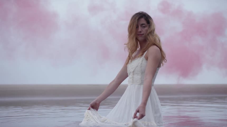 A woman standing in water with a white dress with pink smoke in the background
