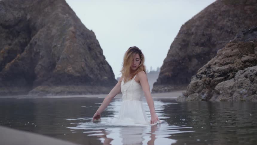 A woman wearing a white dress stands in a lake and plays with the water surrounding her