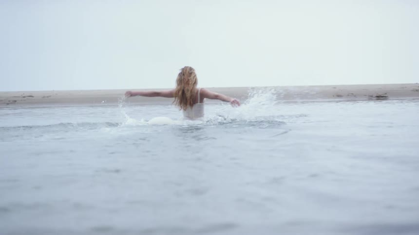 A woman with blonde hair and wearing a white dress runs into a body of water while splashing at it with her hands