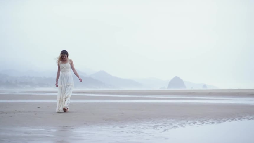 Woman walks across beach barefoot wearing long thin white sundress