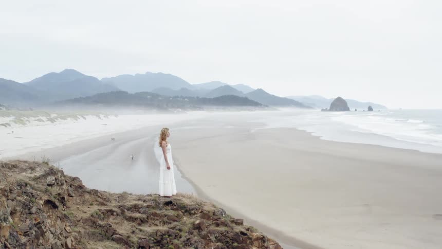 A woman stands a top a tall mountain beside an ocean and beach