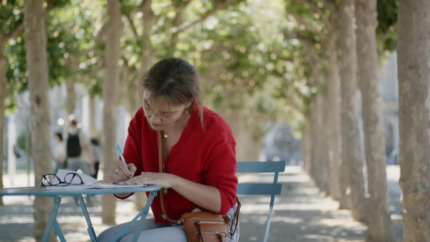 A woman writes at a bistro table under trees