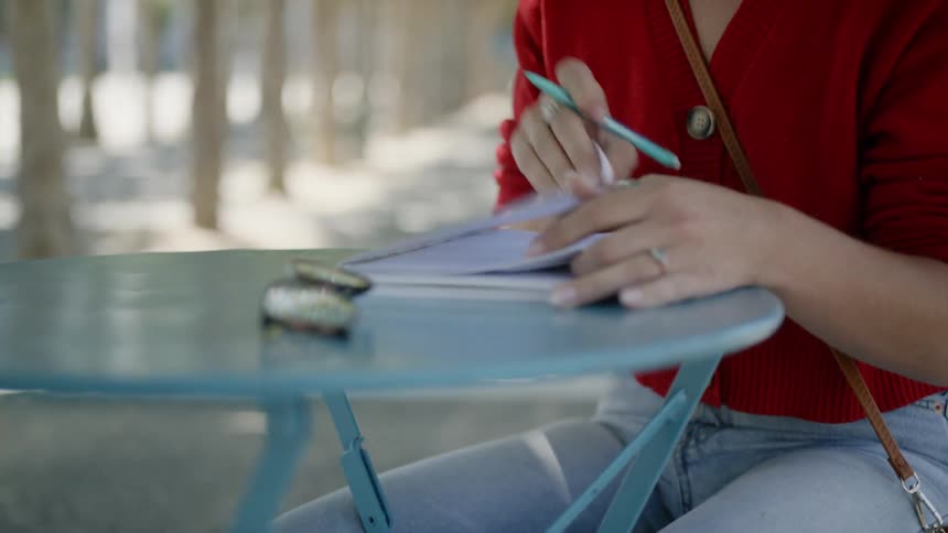 A woman sits at a bistro table writing in a notepad in a park