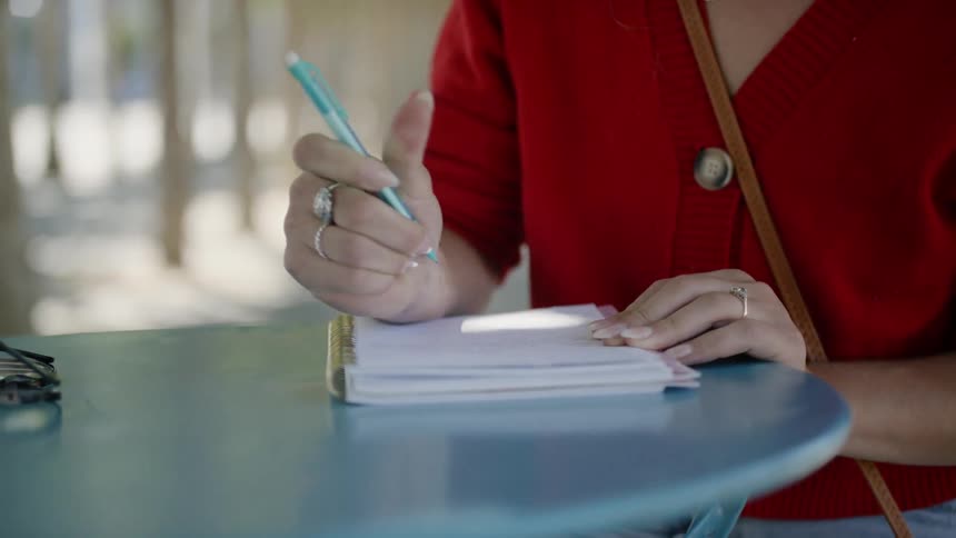 A young woman sits at a table outdoors while writing in a spiral bound notebook