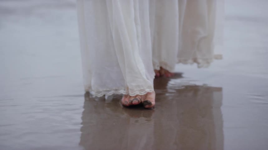 Woman walks barefoot along wet beach in long white skirt