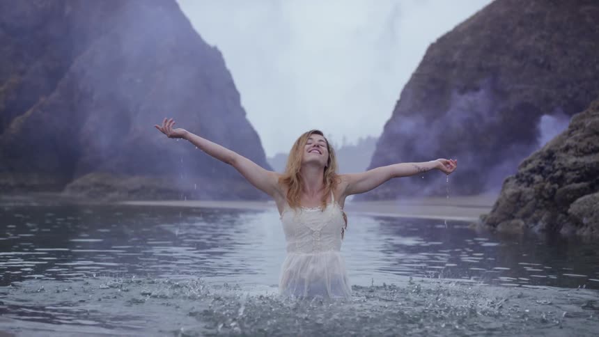 A woman with a white dress standing in water on the beach splashing water and smiling