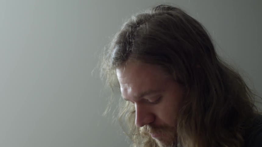 A man with long, blonde hair chewing on a paint brush before he removes it from his mouth, looks down and then back up before placing the brush in his mouth again and walking away