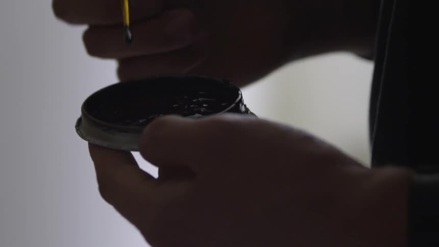 A man dabs a small brush into a small palette of dark paint before lifting the brush