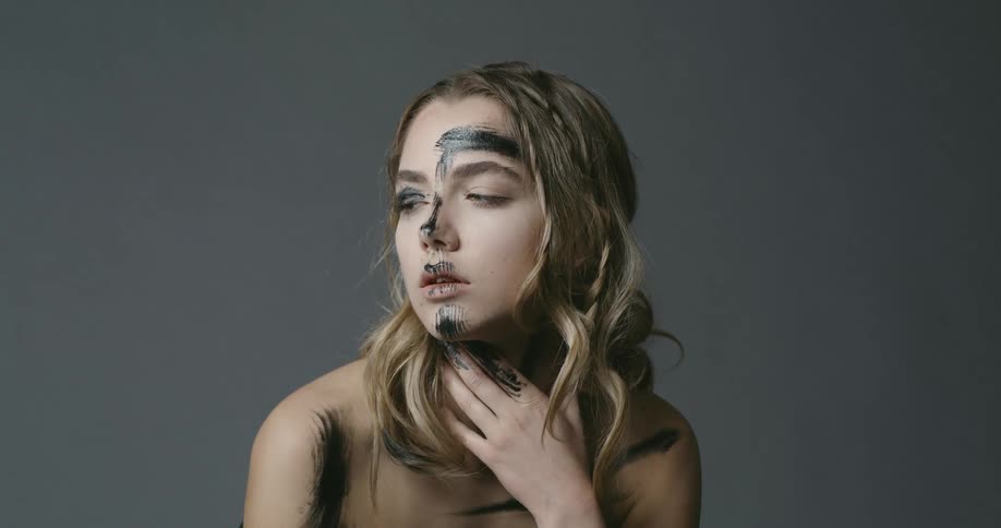 A young woman with black paint smeared across her face looks around sleepily