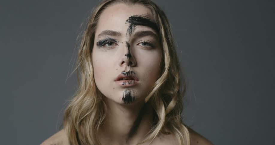 Blonde woman with black brush strokes on her face looks at the camera with a faraway look in his eyes in the studio