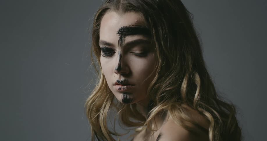 Blonde woman with black brush strokes on her face tilts head down