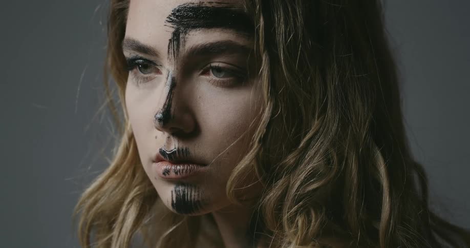 A young woman with streaks of black paint on her face looks downward before briefly glancing up