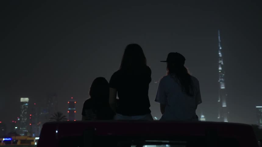 Young women sitting on a jeep looking at a brightly lit city skyline at night and talking