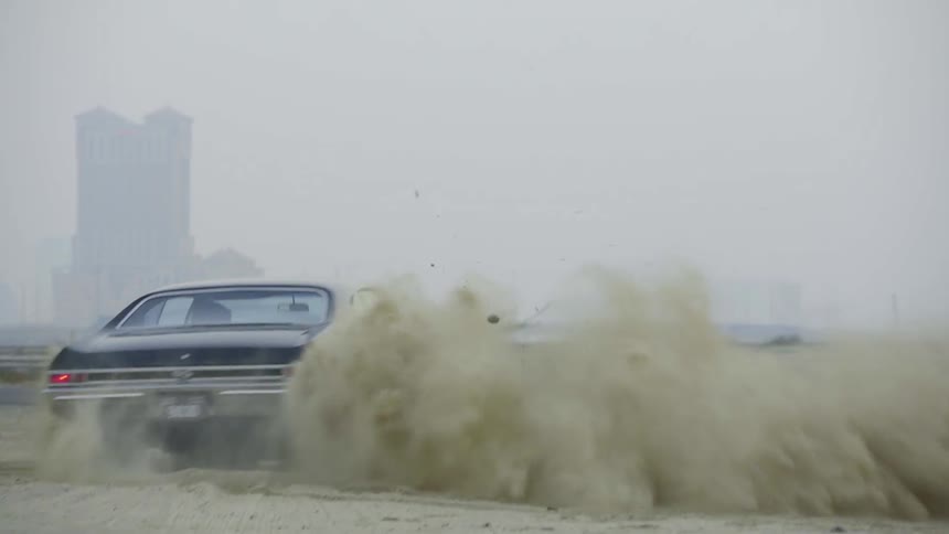 An older black car throwing up dust at a beach
