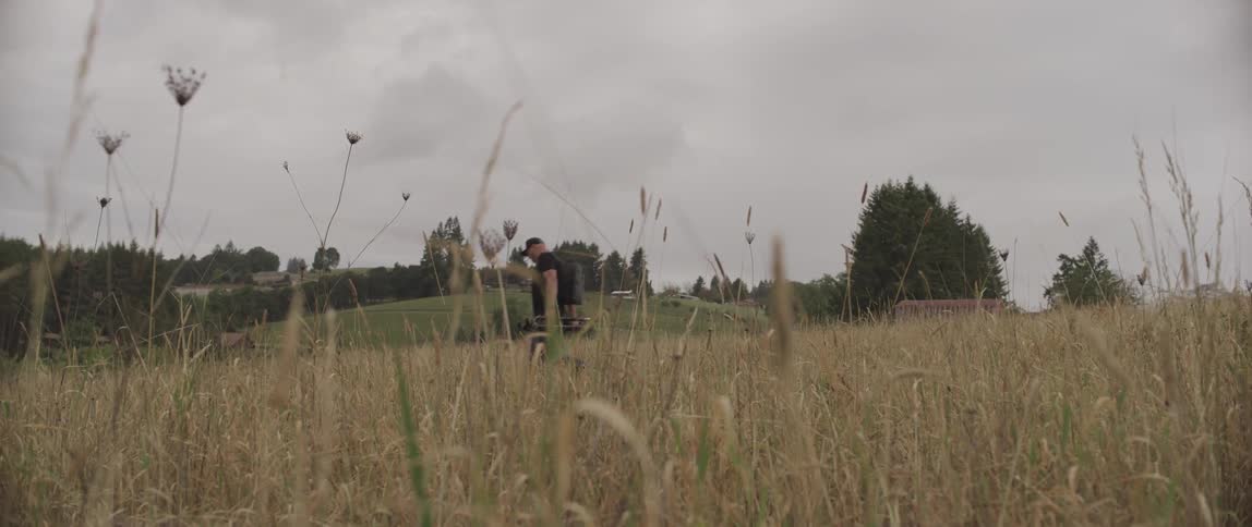 A man walking through a field carrying a tripod on a cloudy day