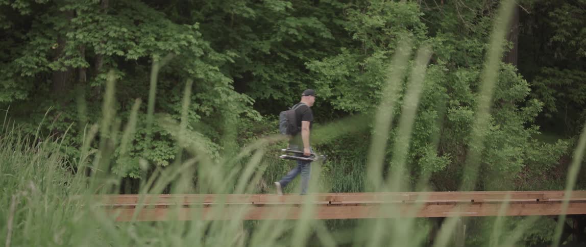 A man with a backpack walks across a bridge carrying a tripod