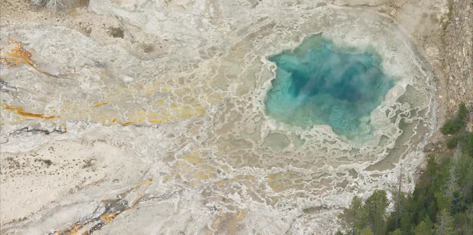 Vapor rises from large blue colored geothermal pool near forest