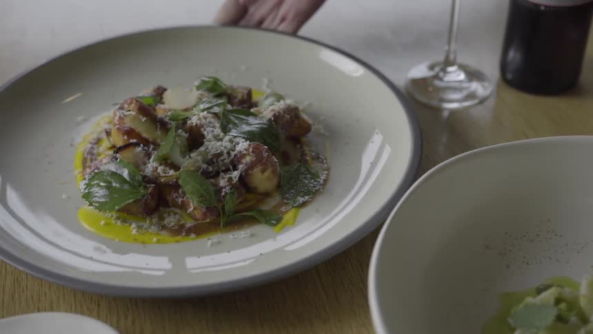 Someone setting a plate of food on a table next to other plates, a wine glass and a bottle