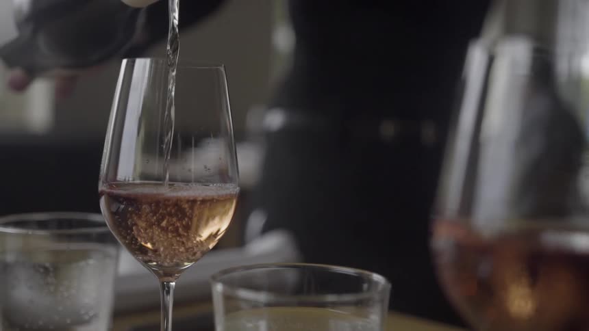 Wine is poured into a tall wine glass that sits on a dining table with place settings