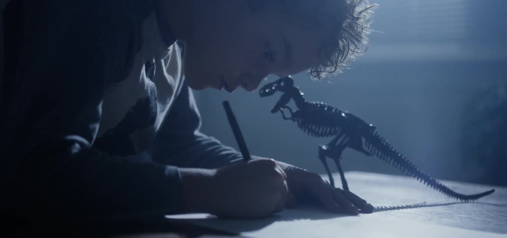 Child draws on table in darkened room next to model of dinosaur skeleton